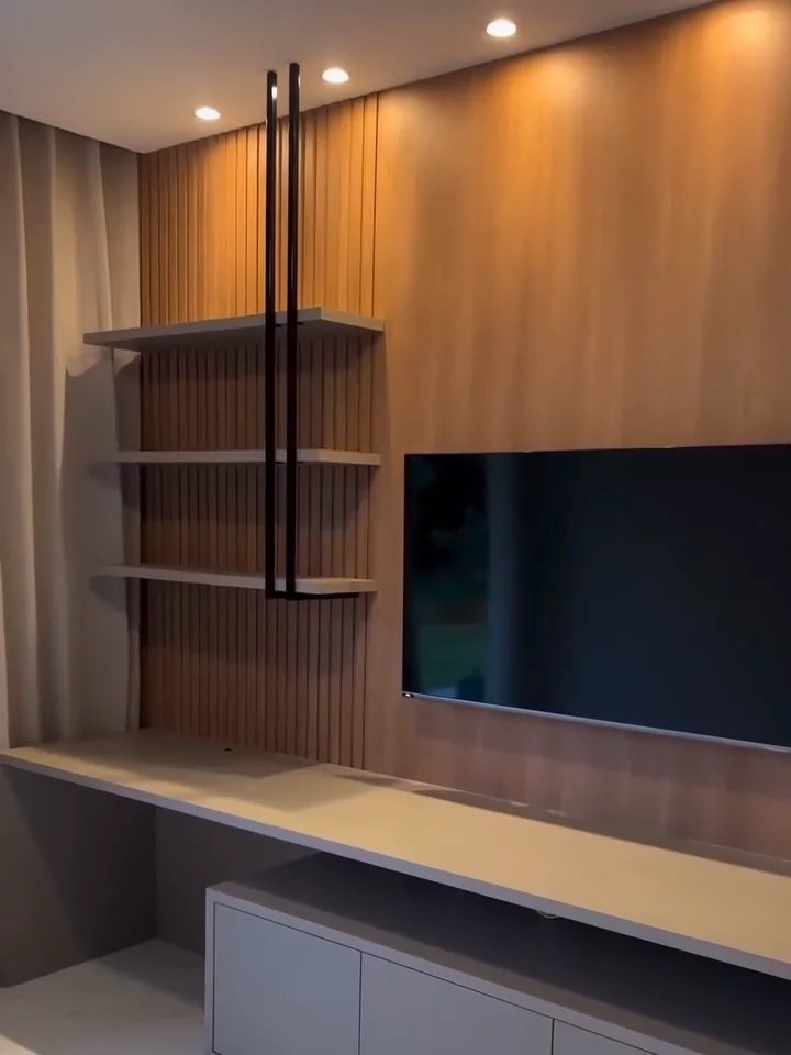 Sala com painel de TV em MDF amadeirado, trecho ripado à esquerda, três prateleiras claras presas por hastes metálicas pretas que descem do teto e rack cinza-claro com tampo comprido